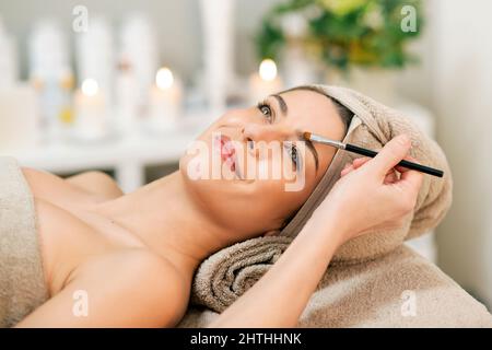 Crop female beauty master with brush applying eyebrow shadow on face of smiling woman in towel lying on couch in salon during relaxing therapy session Stock Photo