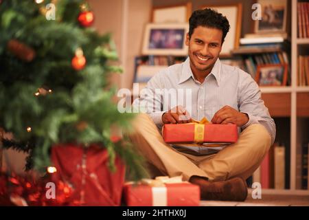Everyone feels like a kid at christmas time. Portrait of a young man sitting next to the tree opening christmas presents. Stock Photo