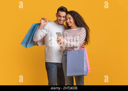 Shopping App. Middle Eastern Spouses Holding Shopper Bags And Using Smartphone Stock Photo
