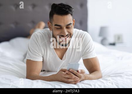 Portrait Of Handsome Young Arab Man Resting In Bed With Smartphone Stock Photo