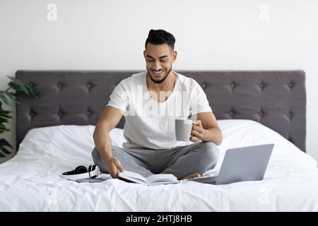 Portrait Of Arab Man Sitting In Bed With Laptop, Notepad And Coffee Stock Photo
