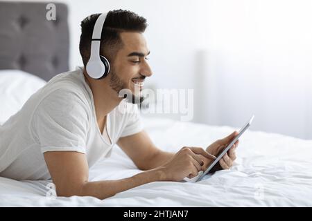 Cheerful Young Arab Man Relaxing With Digital Tablet In Bed Stock Photo