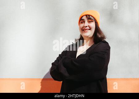 Young smiling female with green dyed hair wearing a beanie and a black jacket looking at camera while standing near orange and grey wall on sunny stre Stock Photo