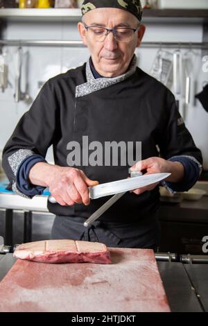 Mature male chef in uniform sharpening blade of knife with honing steel while getting ready for cooking beef in kitchen of restaurant Stock Photo