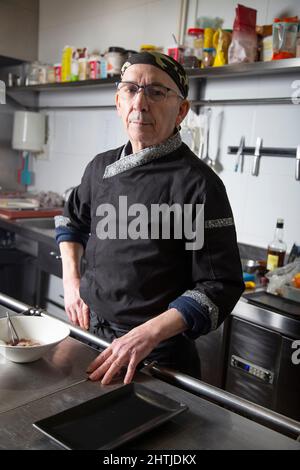 Self assured mature male cook in uniform standing at stainless steel table in kitchen of restaurant and looking at camera Stock Photo