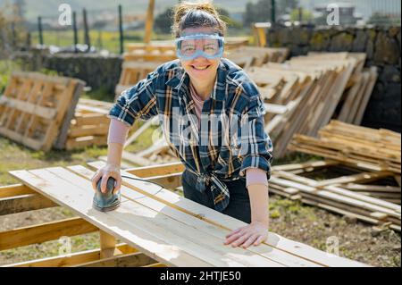 Cheerful female artisan in protective glasses using special electric orbital sander to smoothen wooden planks while working in countryside on summer d Stock Photo