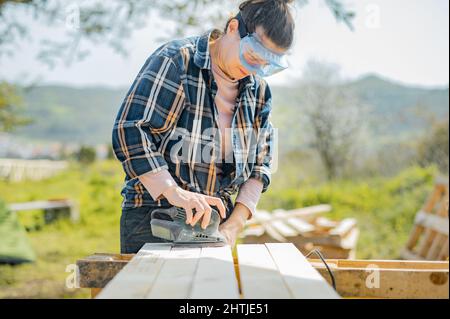 Focused female artisan in protective glasses using special electric orbital sander to smoothen wooden planks while working in countryside on summer da Stock Photo