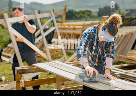 Focused female sanding down wooden planks with special electric orbit sander while working in countryside near man carrying wooden construction Stock Photo