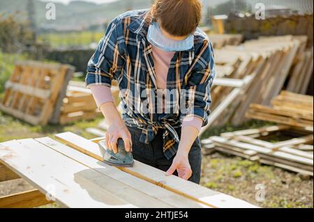 Anonymous female artisan in protective glasses using special electric orbital sander to smoothen wooden planks while working in countryside on summer Stock Photo