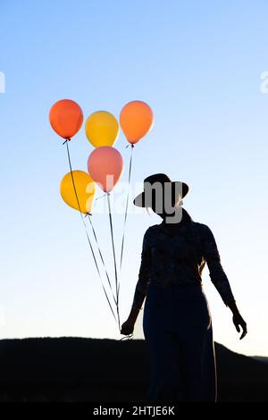 Silhouette of unrecognizable female in hat standing with heap of colorful balloons against cloudless sky at sunset time against mountain ridge Stock Photo