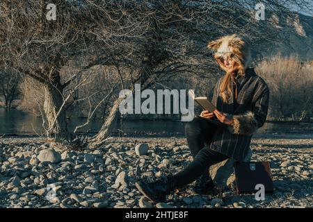 Woman in coat and fox fur hat sitting on stony coast of lake and reading book in fall day Stock Photo