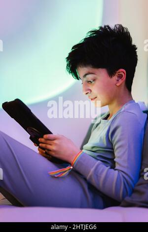 Side view of barefoot Arab teen boy sitting on bed and browsing Internet on tablet in bedroom Stock Photo