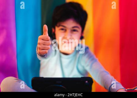 Positive Arab boy in wireless earbuds sitting near colorful rainbow flag and watching online video on tablet while showing like gesture and looking at Stock Photo