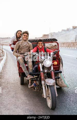 Luxor, Egypt - December 25, 2021: Delightful kids riding vehicle on asphalt road while collecting rubbish for living Stock Photo