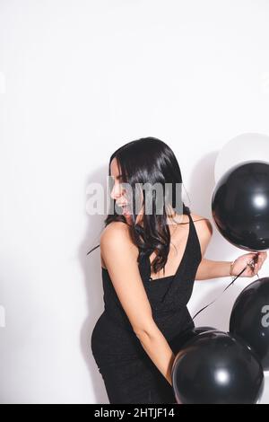 Charming female in stylish black dress with bunch of balloons in hand smiling while celebrating birthday on white background Stock Photo