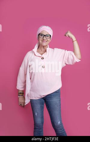 Positive mature female with cancer wearing headscarf and showing bicep while looking at camera on pink background in light studio Stock Photo