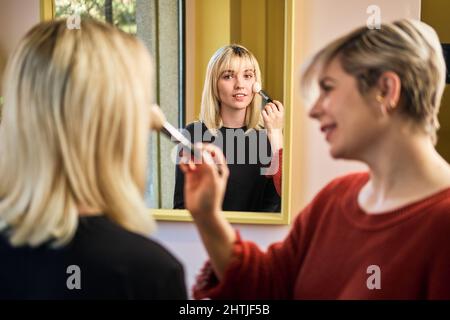 Positive female visagiste applying powder on face of young attractive Woman with brush while sitting in salon with mirror during beauty procedure Stock Photo