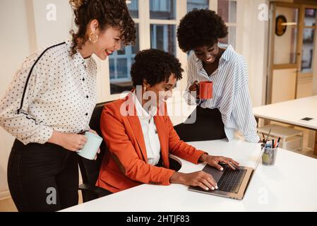 Group of happy multiethnic female coworkers in elegant wear surfing netbook while working together on project in light modern office Stock Photo