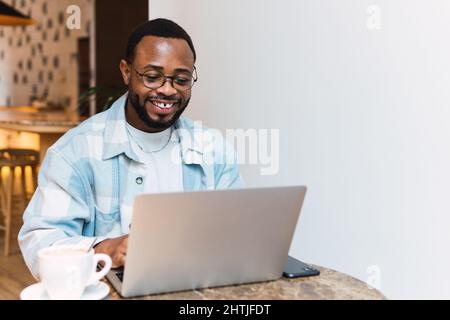 Smiling African American male with cup of coffee surfing netbook at table in modern cafe during break Stock Photo