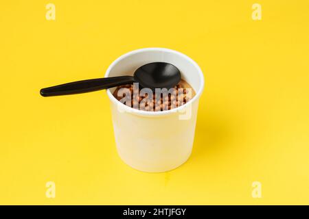 White paper tub filled with sweet chocolate ice cream topped with small candies placed on yellow background with black spoon Stock Photo