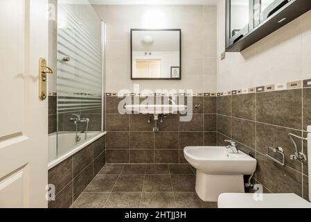 Large bathroom with separate shower, white porcelain toilets, tile trim, gray tile and brown framed mirror and black cabinets Stock Photo