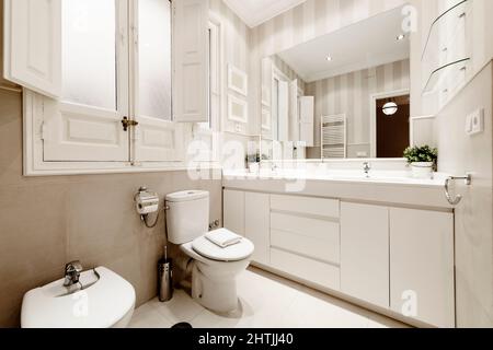 One-piece white porcelain washbasin with two bowls on a white wooden cabinet with a square frameless mirror and a white heated towel rail next to the Stock Photo