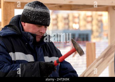 A builder in a blue jacket with a hammer in his hand hammers a nail Stock Photo