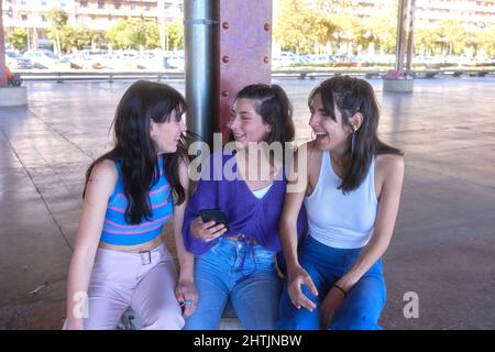 Three friends laughing while using the mobile sitting on an urban area Stock Photo