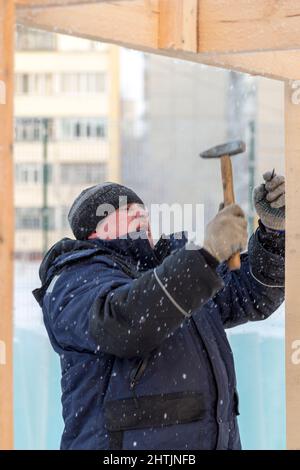 A builder in a blue jacket with a hammer in his hand hammers a nail Stock Photo