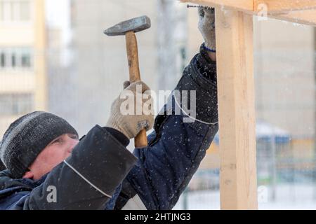 A builder in a blue jacket with a hammer in his hand hammers a nail Stock Photo