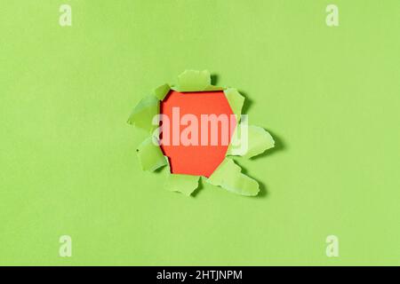 A hole in green paper with torn edges and a red  background inside. Ripped green paper, copy space Stock Photo