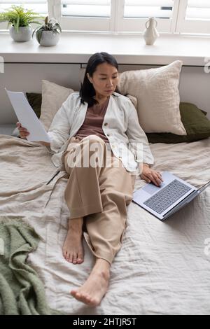Young Asian female auditor with papers sitting on double bed and looking through online data about business development Stock Photo