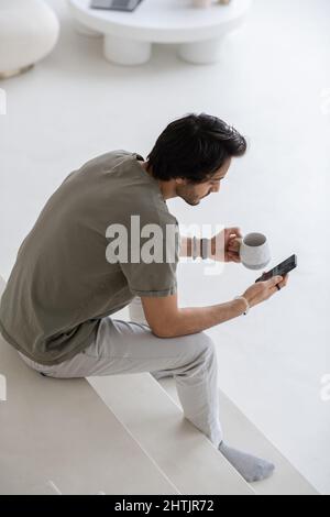 Young modern businessman with mobile phone sitting on white staircase in the morning and texting or scrolling through online news Stock Photo