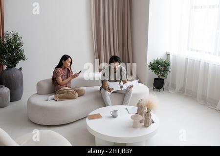 Young Caucasian businessman reading financial papers while sitting on sofa next to Asian female texting in smartphone Stock Photo