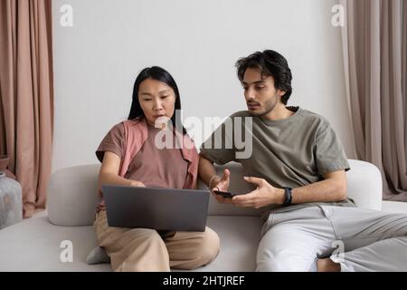 Two intercultural employees discussing online information on laptop screen while Asian female explaining points of presentation Stock Photo