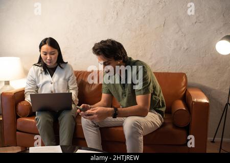 Two young interracial business people using wireless gadgets while sitting on brown leather sofa in living room Stock Photo