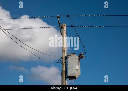 Simple transformer connection on a wooden pylon electricity pole carrying two cables, blue sky and fluffy white cloud behind Stock Photo