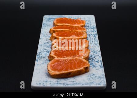Traditional Japanese Tataki, slices of wild and fresh salmon of norway cooked outside and raw inside served in ceramic plate on black background. Stock Photo