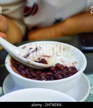 Hand spoons up red bean double skin milk pudding from a white bowl. Double skin milk pudding is a popular dessert in Hong Kong. Stock Photo