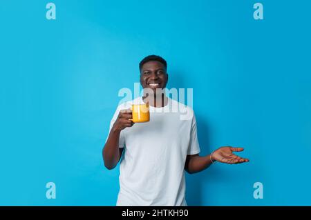 An image of a dark-skinned man in a white T-shirt with a yellow cup of coffee in his hand on a blue background. Stock Photo