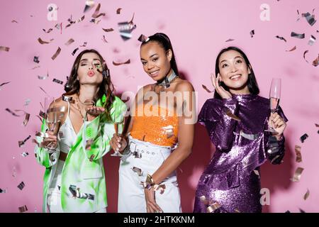happy multiethnic women holding glasses of champagne near model showing air kiss near falling confetti on pink Stock Photo