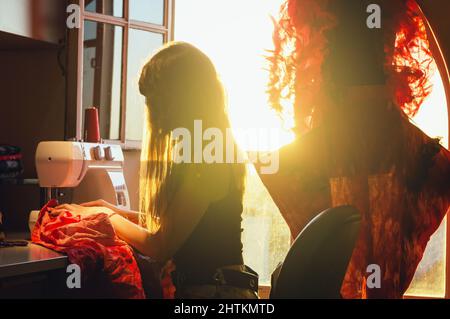 Empowered enterprising latin caucasian young woman working in her sewing workshop, sitting by the window lit by sunlight using the sewing machine sewi Stock Photo