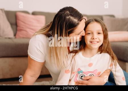 Close up photo of two people mother and her little nice daughter sitting on the carpet at home and mother hugging and kissing her little girl on the Stock Photo