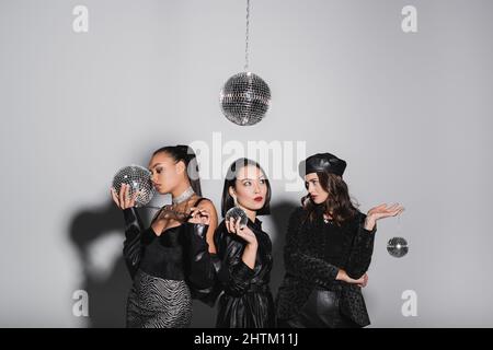 young woman in beret posing near interracial models holding disco balls on grey Stock Photo