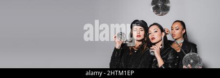 young woman in beret posing near interracial models holding disco balls on grey, banner Stock Photo