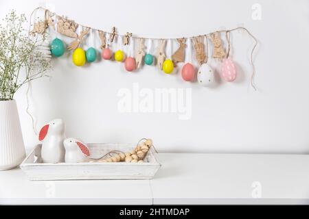 Fragment of the interior. Decorated children's room for Easter. A garland of plastic eggs on the wall. Ceramic rabbits and wooden cubes on the table. Stock Photo