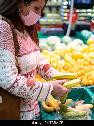 Middle-aged woman with a pink face mask holds bananas in her hands in a supermarket. She examines the bananas to select the best ones to buy. Stock Photo