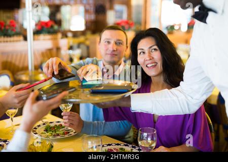 African american waiter collecting phones of cheerful company in restaurant Stock Photo