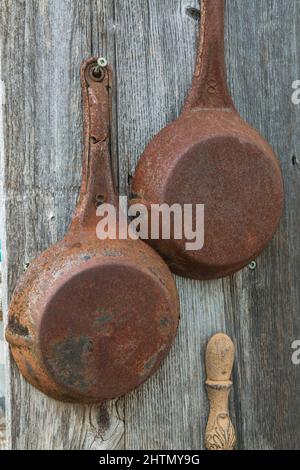 Old rusted frying pans hanging on grey weathered exterior wall of old rustic wood plank cabin. Stock Photo