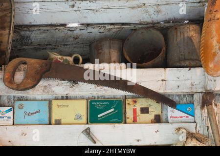 Antique saw, empty paint cans, metal cigarette boxes on shelf inside old rustic wood plank cabin. Stock Photo